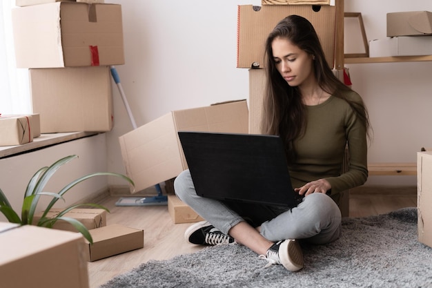 chica feliz con cajas se muda a un nuevo apartamento se muda a una nueva casa alquiler de viviendas española mujer joven usando computadora portátil para buscar y pedir un servicio de transporte y se muda a unha nueva casa