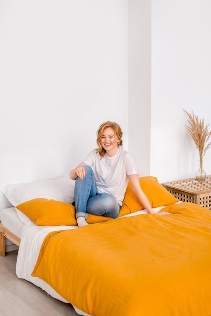 Chica feliz en una camiseta blanca y jeans azules está sentada en la cama Buenos días