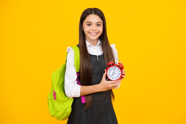 Chica feliz cara positiva y sonriente colegiala adolescente con mochila mantenga la alarma del reloj para aprender Colegiala sobre fondo amarillo aislado