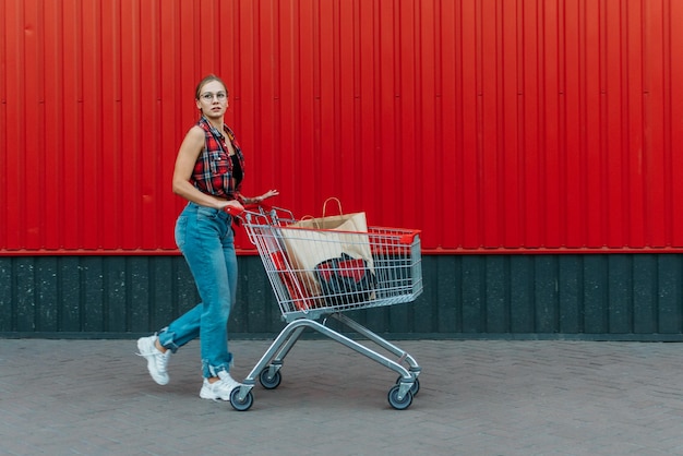 Foto chica feliz con un carrito de compras en el fondo de la tienda de la pared roja mujer joven empujando un carrillo de compras