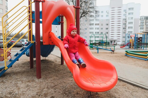 Chica feliz en una chaqueta roja cabalga sobre una colina en el patio de recreo divertida niña alegre jugando