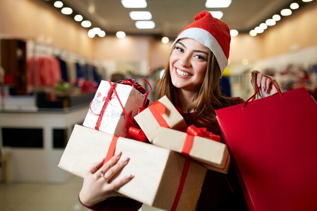 Foto chica feliz comprando regalos en el centro comercial en venta de navidad concepto de idea de compras de vacaciones de año nuevo mujer sonriente con papel de colores presenta bolsas y cajas de regalo con sombrero de navidad en la tienda o tienda