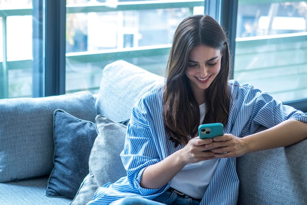 Foto chica feliz desplazándose y revisando las redes sociales mientras sostiene el teléfono inteligente en casa sonriendo joven mujer morena usando una aplicación de teléfono móvil para jugar juegos tienda entrega de pedidos en línea mientras se relaja en el sofá