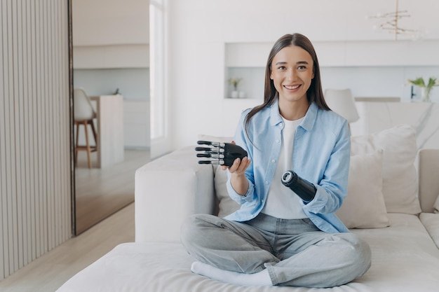 Chica feliz con discapacidad disfrutando usando prótesis biónica de brazo sentado en el sofá en casa