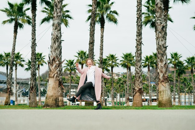 Chica feliz y elegante con un largo abrigo rosa caminando en un parque verde junto a palmeras altas