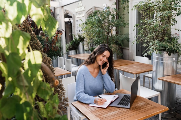 La chica feliz está hablando por teléfono y conversando con sus colegas de forma remota. La gerente alegre con una computadora portátil está tomando notas en el cuaderno y hablando con sus colegas.