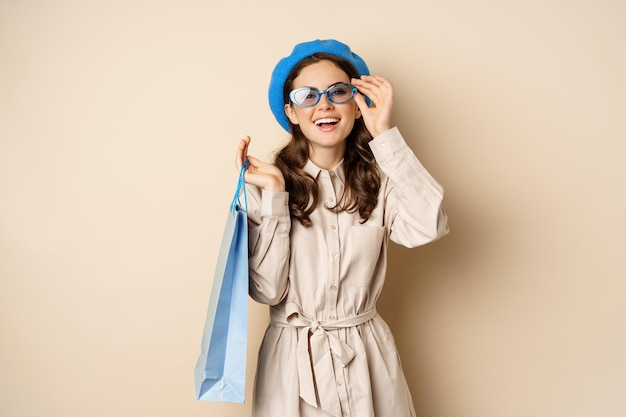 Chica feliz con estilo en las compras. Retrato de una mujer moderna con una bolsa de compras, riendo y sonriendo satisfecha, comprándose un regalo, de pie sobre un fondo beige