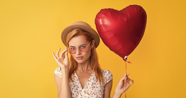 Una chica feliz con gafas de sol sostiene un globo de corazón de amor con fondo amarillo