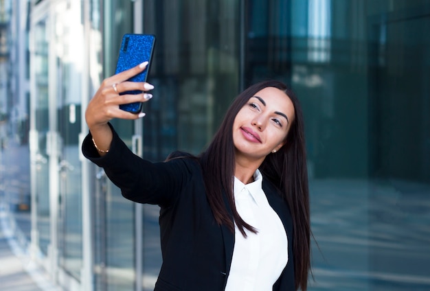 Chica feliz glamorosa con grandes labios y uñas largas o joven empresaria tomando selfie en su teléfono celular. Formal vestida alegre mujer en un traje haciendo una llamada de video, sonriendo, mirando el teléfono inteligente.