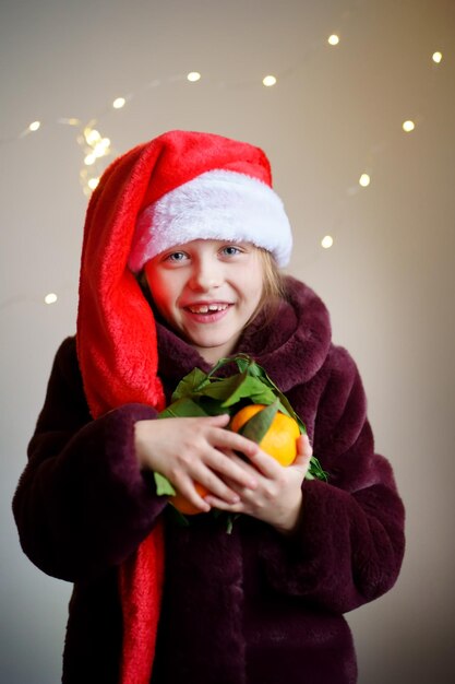 Una chica feliz con una gorra de Santa y un abrigo de piel tiene un montón de mandarinas hay luces de fondo