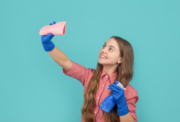 Chica feliz en guantes de goma con botella de spray y trapo de microfibra sobre fondo azul.