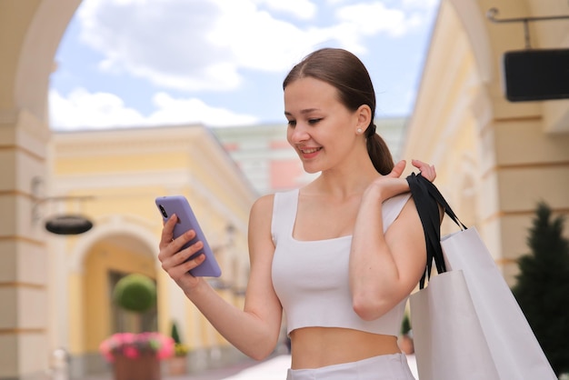 Chica feliz joven es una sonrisa mirando la pantalla del teléfono inteligente en el centro comercial que lleva una bolsa de papel de compras