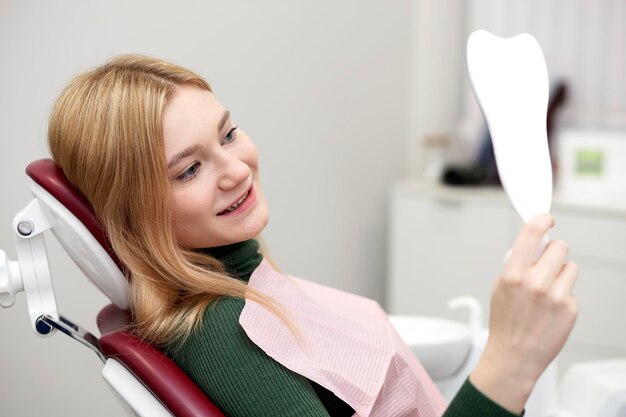 Chica feliz joven mujer sonriente satisfecha paciente con una sonrisa perfecta mirando al espejo después del dentista