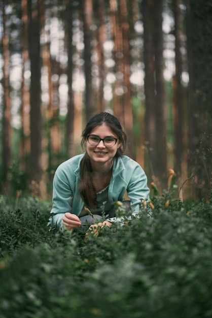 Chica feliz lleva gafas recogiendo bayas en el bosque mirando a la cámara Retrato de una mujer sentada en los arbustos sonriendo a la cámara