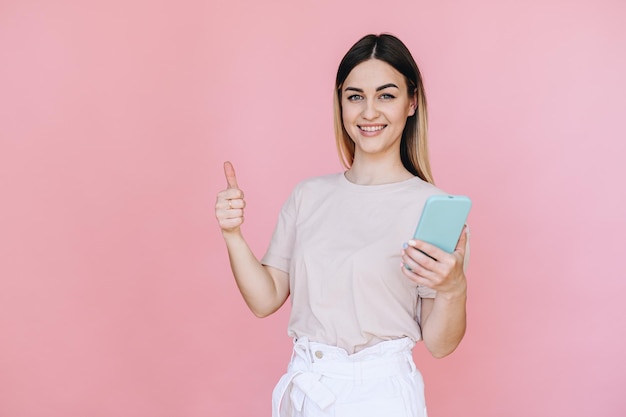 Chica feliz mostrando los pulgares hacia arriba y sosteniendo un teléfono en la mano sobre un fondo rosa