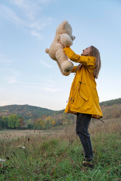 Chica feliz con osito de peluche en chaqueta amarilla del parque se encuentra en el fondo de las montañas Mujer joven con osito de pelucheVertical