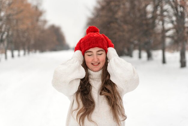 Chica feliz riendo con los ojos cerrados en sombrero rojo y mitones en el parque de invierno Hermosa mujer joven en caminar de invierno
