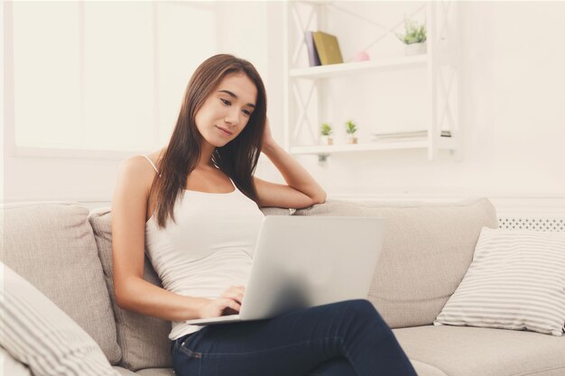 Chica feliz sentada cómodamente en un sofá beige usando una laptop, enviando mensajes de texto en línea. copia espacio