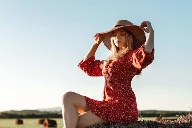 Foto chica feliz sentada en un puñado de heno en un campo de verano al atardecer