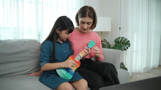 Chica feliz tocando el ukulele mientras la madre caucásica enseña y explica la pedagogía