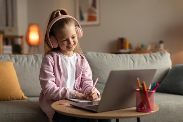 Chica feliz usando una computadora portátil tomando notas usando auriculares en casa