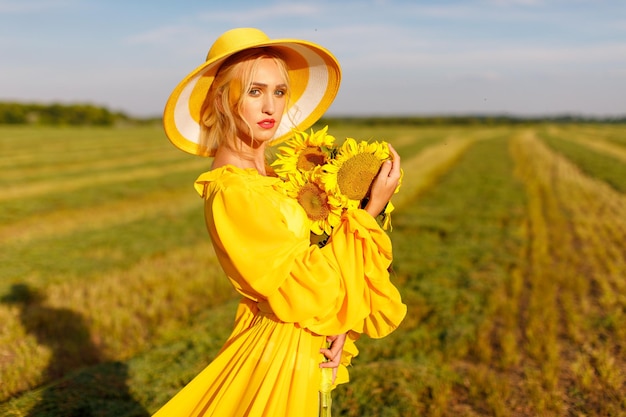 chica feliz con un vestido amarillo sostiene flores de girasol en un campo de girasoles contra el cielo