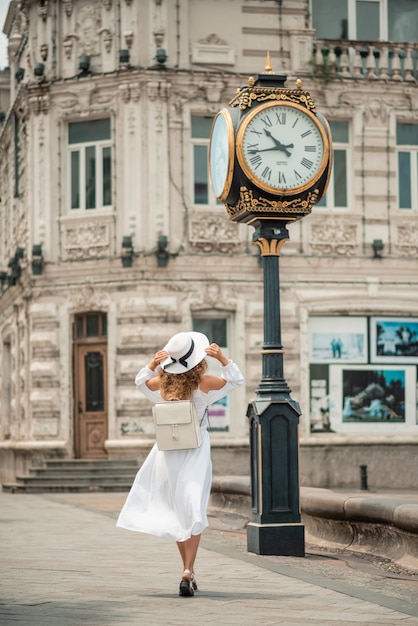 Chica femenina en un vestido largo blanco, con una mochila, sosteniendo el sombrero caminando cerca del reloj