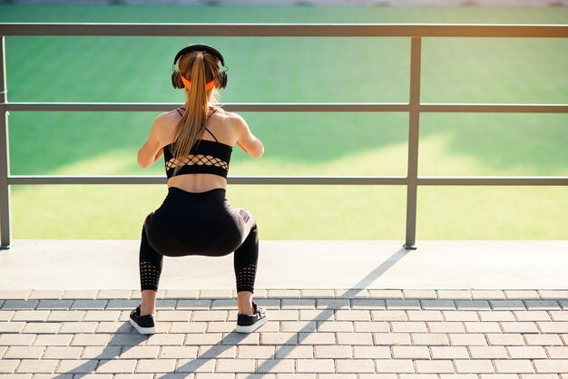 Chica fitness con auriculares que hace ejercicios de sentadillas con una banda de botín de tela durante su entrenamiento deportivo en un campo deportivo especial