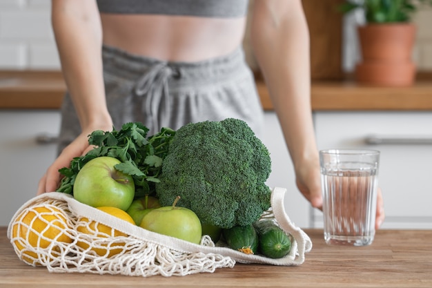 Chica fitness irreconocible con bolsa de malla llena de frutas y verduras saludables en el fondo de la cocina moderna