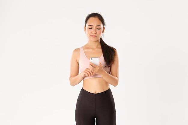 Chica fitness mirando la pantalla de su teléfono inteligente con cara de malestar y sonrisa, decepcionada con los resultados de ejecución de la aplicación de seguimiento de entrenamiento.