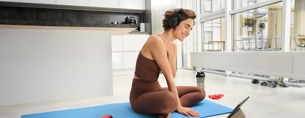 Una chica de fitness morena sonriente escucha música en los auriculares ve videos en línea desde la tableta aprende