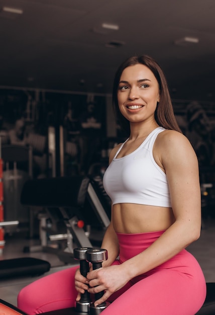 Chica fitness con pesas posando en un banco en el gimnasio