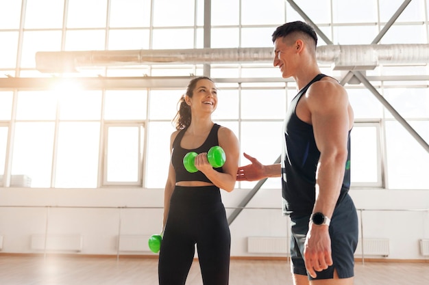 Una chica de fitness en ropa deportiva se entrena con un entrenador en un gimnasio brillante. Un entrenador masculino ayuda a una mujer a levantar pesas.