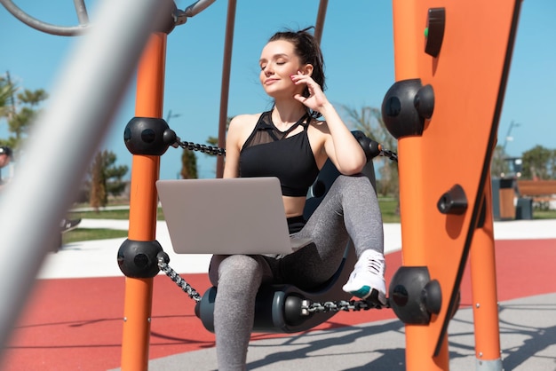 Chica fitness en ropa deportiva sentada en el patio de recreo en verano al aire libre con una computadora portátil