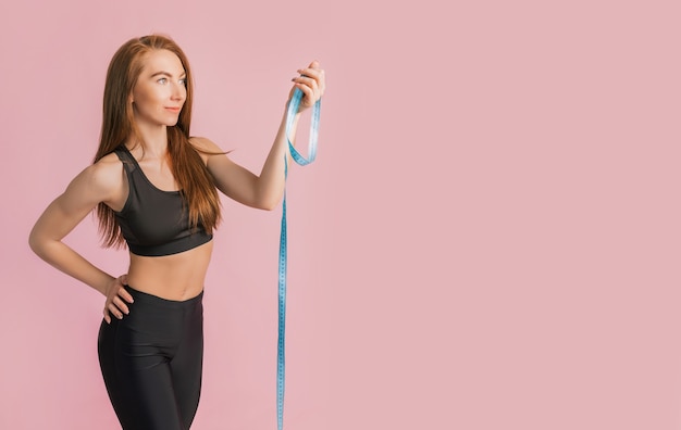 Chica fitness sonriendo y posando mantenga cinta métrica en ropa deportiva negra en un espacio rosa