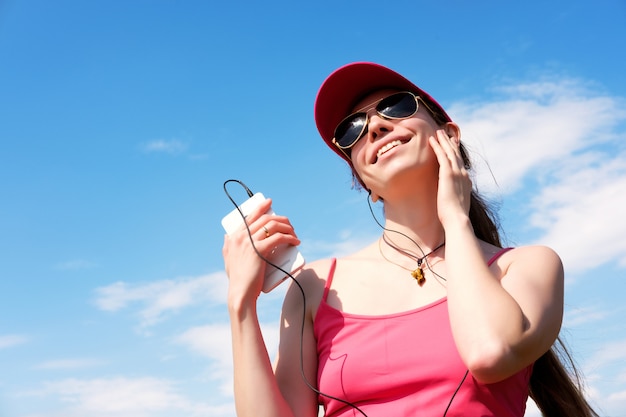 Chica flaca pelirroja con gorra rosa y gafas de sol está escuchando música durante trotar en verano, copia espacio