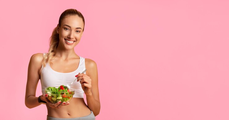 Foto chica en forma comiendo ensalada de verduras de un cuenco sonriendo a la cámara de pie sobre el fondo amarillo del estudio