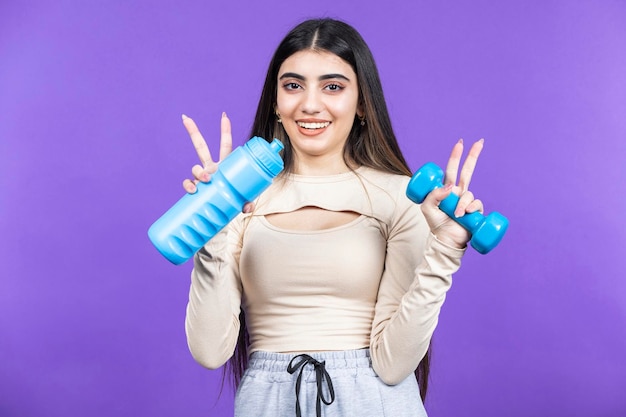 Chica en forma sonriente sosteniendo una botella de agua y mancuernas Chica en forma joven de pie sobre fondo púrpura Foto de alta calidad