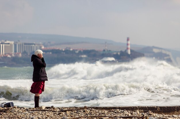 Chica fotografía grandes olas de tormenta junto a un faro y rocas. La niña está vestida con una chaqueta negra, gorro de punto blanco, falda larga burdeos y botas negras.