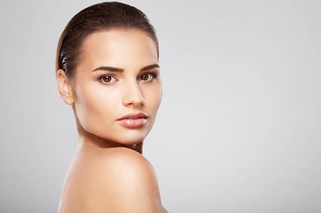 Chica fresca con cabello castaño, piel limpia y fresca y hombros desnudos posando en el fondo gris del estudio, un modelo con un ligero maquillaje nude, de cerca.