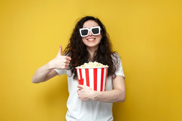 Chica con gafas 3d sostiene palomitas de maíz, mira una película y muestra como con su mano sobre un fondo amarillo