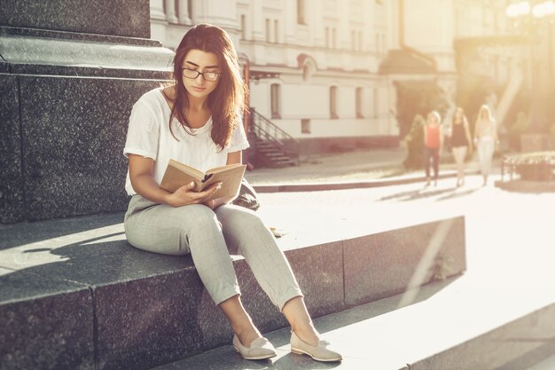 Una chica con gafas lee un libro cerca de la universidad