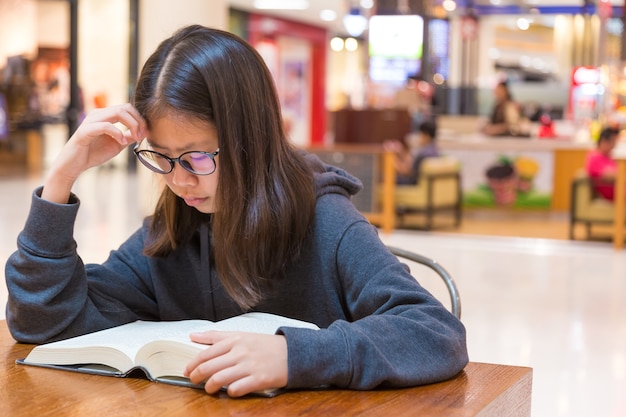 Foto chica con gafas leyendo un grueso libro de cuentos de ficción en un lugar público