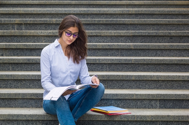 Una chica con gafas con un libro se sienta en los escalones