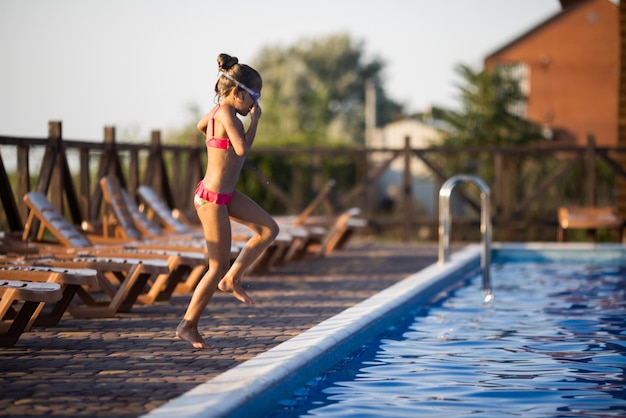 Una chica con gafas de natación salta a una piscina con agua clara en el fondo de una cálida puesta de sol de verano
