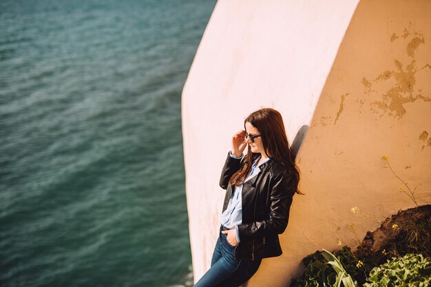 Chica con gafas de sol descansa contra la pared amarilla cerca del mar.