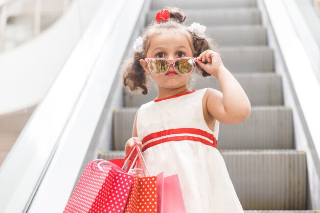 Chica con gafas de sol en una escalera mecánica en un centro comercial con bolsas de colores