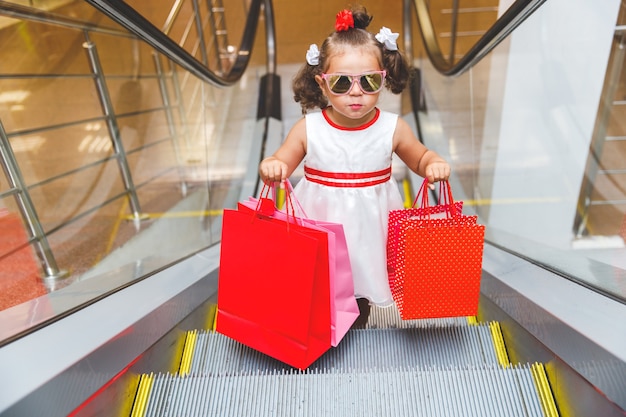 Chica con gafas de sol en la escalera mecánica del centro comercial con compras