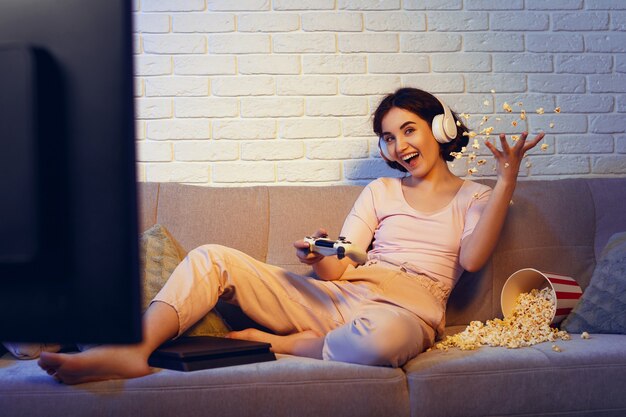 Chica gamer involucrada usando audífonos y jugando videojuegos en la consola en casa. mujer está comiendo palomitas de maíz