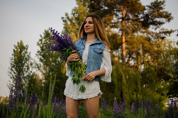 Chica en general beige y chaqueta de jeans de pie en el campo con el ramo de flores